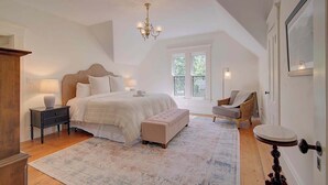 A bedroom with a large bed, a wooden nightstand, a bench, a chair, a chandelier, and a window with natural light. - Short Drive to Gray's Beach! (Yarmouth)
