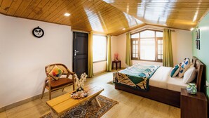 Une pièce avec un plafond en bois, un lit avec une literie à motifs, une chaise avec un coussin coloré, une petite table avec un vase et une fenêtre avec des rideaux.