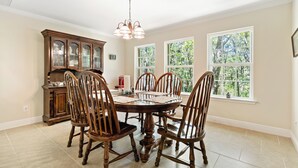 Uma sala de jantar com mesa e cadeiras de madeira, um candelabro e janelas amplas com vista para o verde.