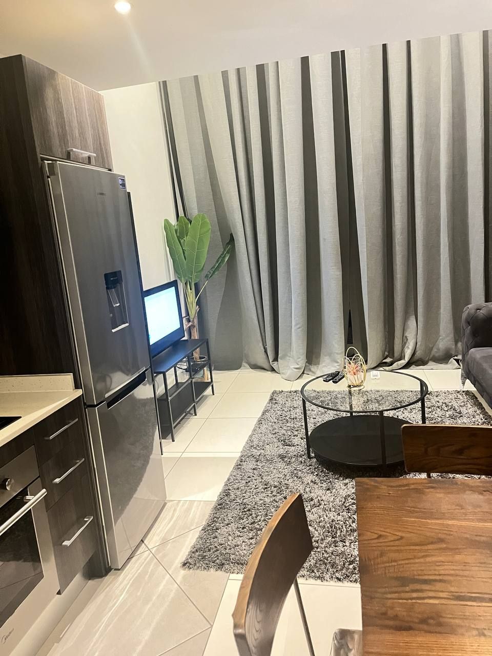 Una sala de estar moderna con área de cocina, que incluye un refrigerador de acero inoxidable, un televisor de pantalla plana, una mesa de comedor con sillas y una mesa de centro.