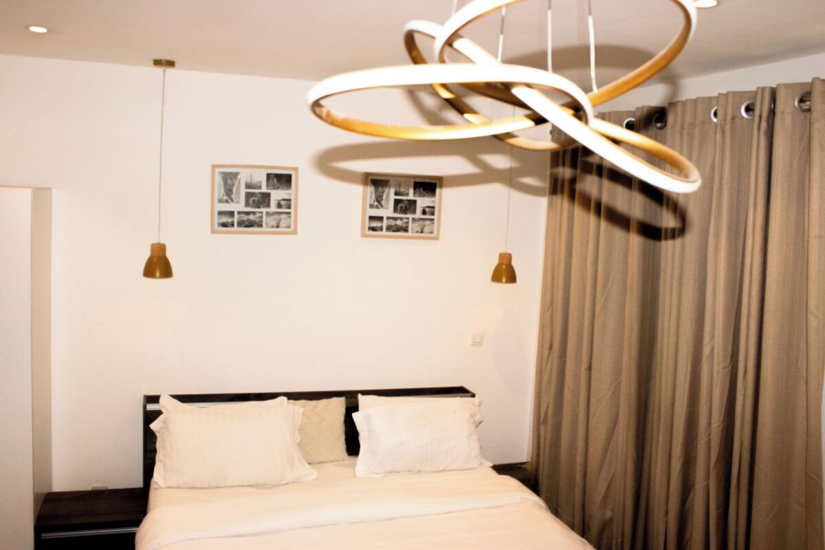 A neatly made bed with white linens and two pillows, a modern circular light fixture above, and two framed pictures on the wall.
