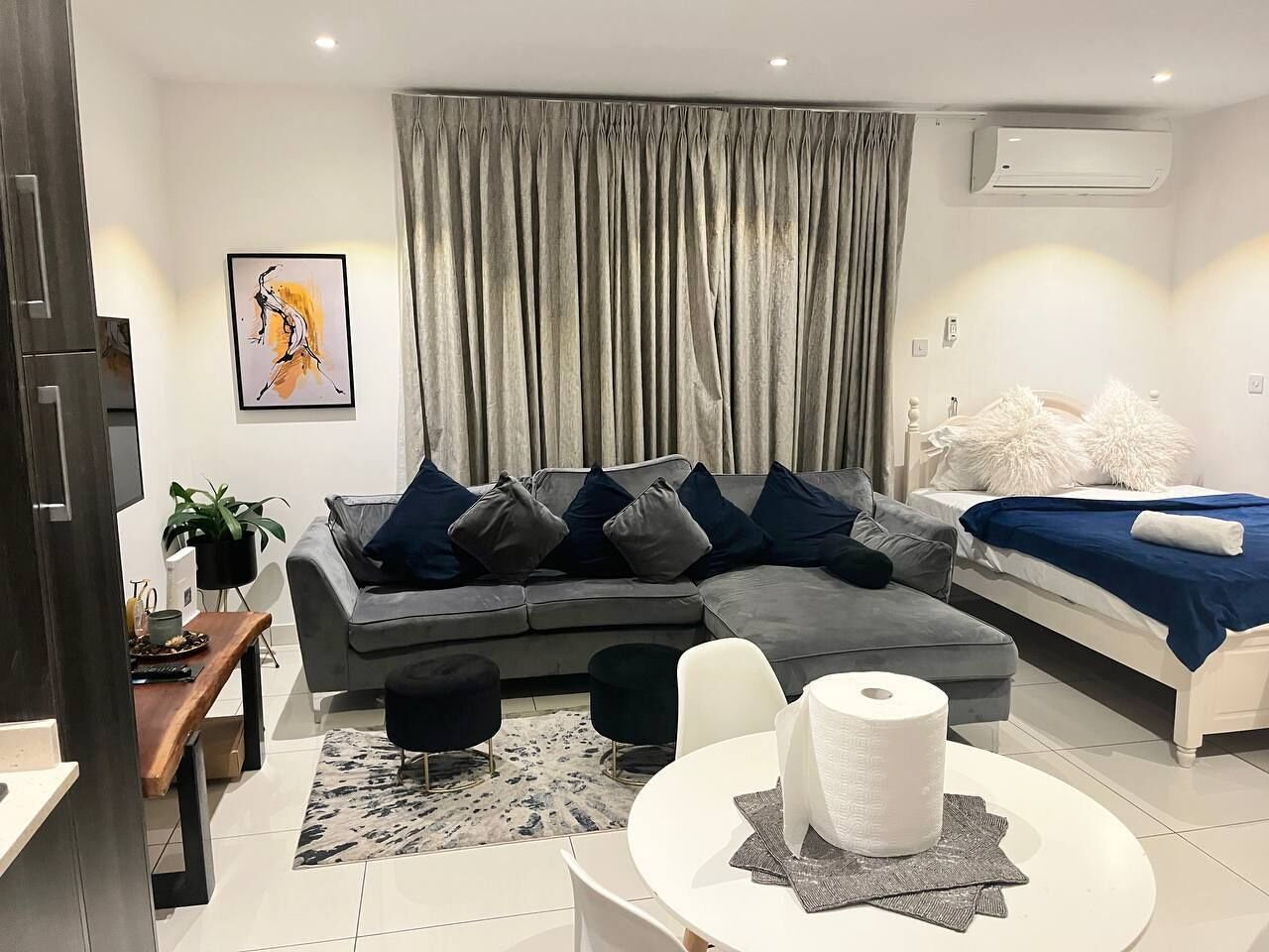 A modern living room with a grey sectional sofa, a white round table with a roll of toilet paper, a grey and white bed with white pillows, and a grey curtain.