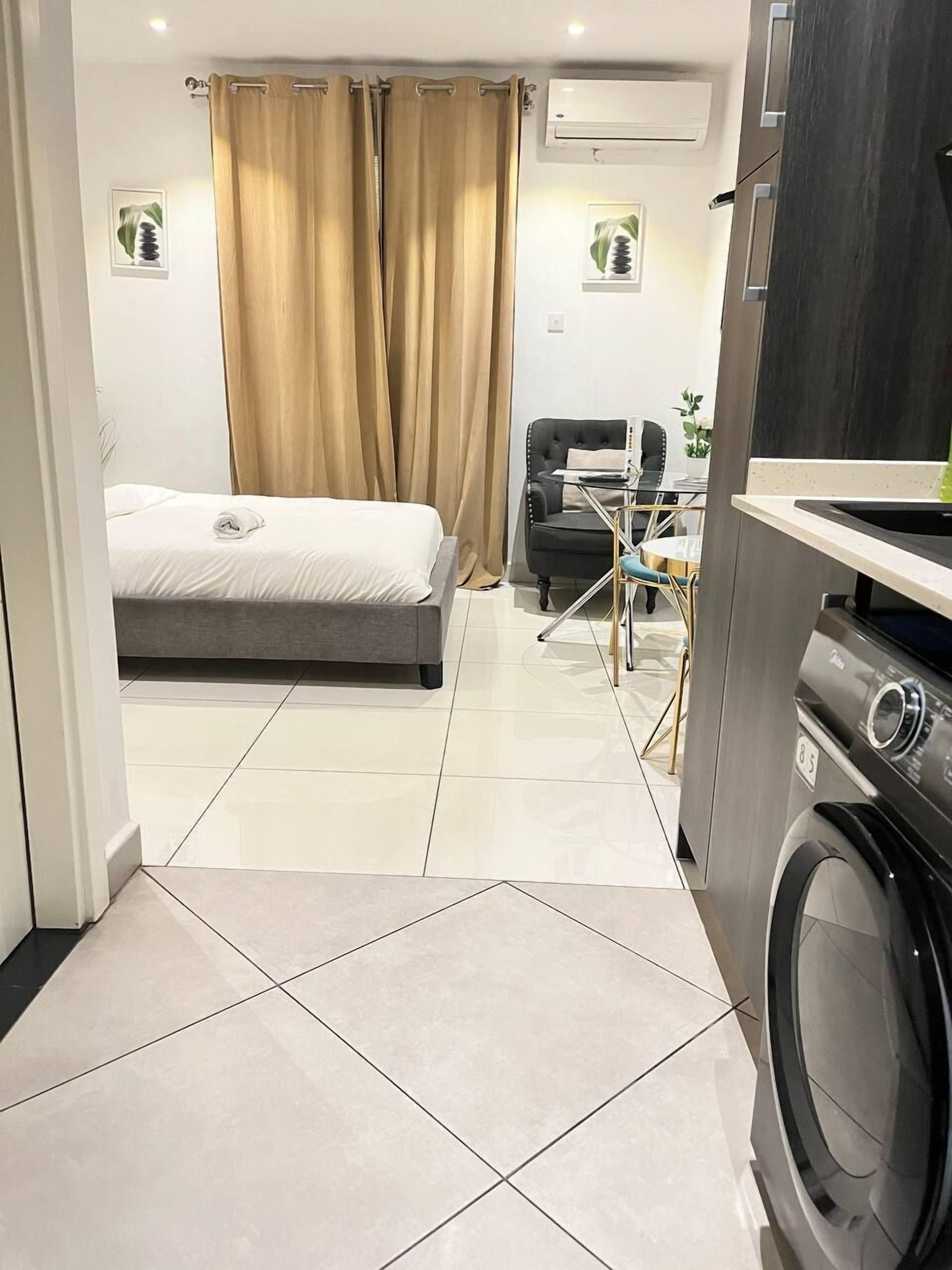 A hotel room with a bed, a chair, a small table, an air conditioning unit, and a washing machine.