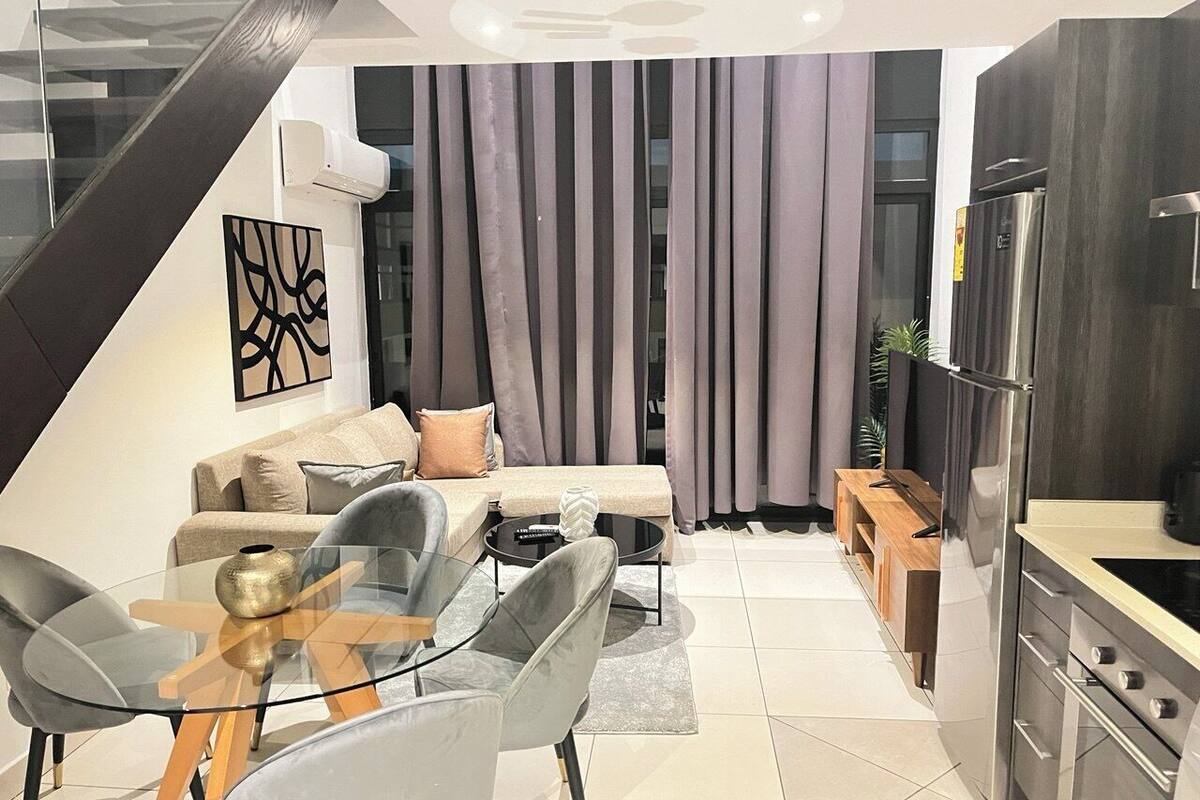 A modern living room with a glass dining table, grey chairs, a sofa set, and a kitchen area with a refrigerator and stove.