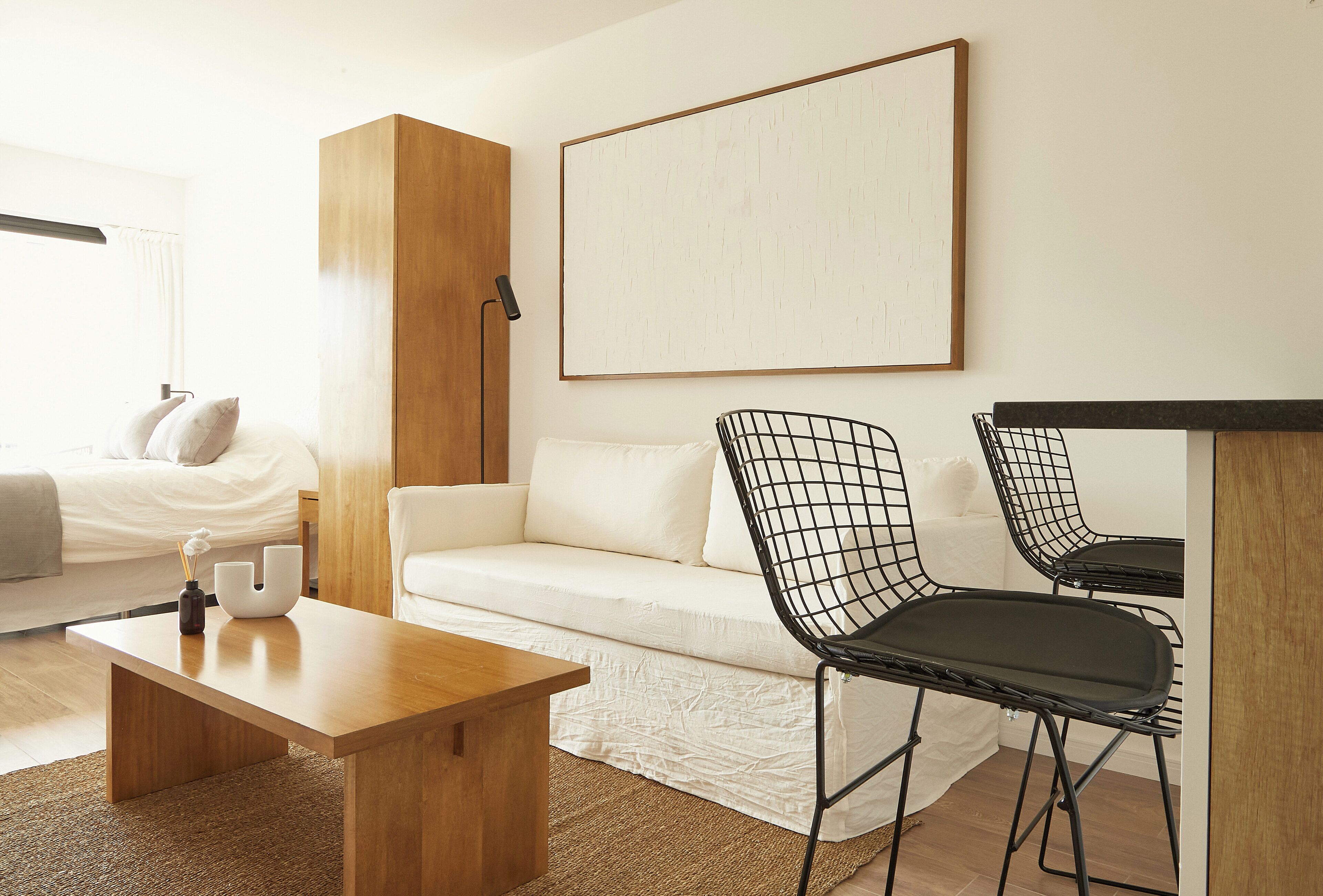A modern, well-lit room with a wooden coffee table, a white sofa, and two black wireframe chairs. A large abstract painting hangs on the wall, and a bed with white bedding is visible in the background.