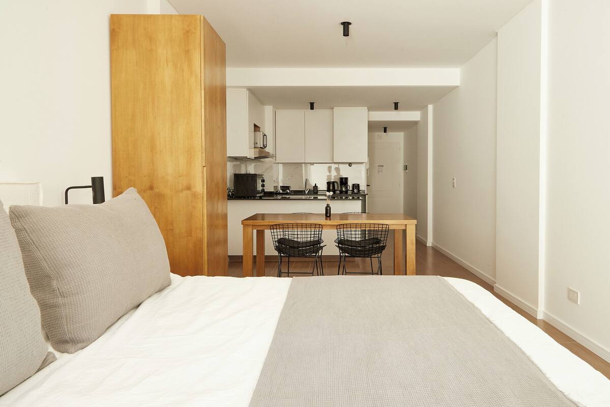 A modern hotel room with a large bed, a wooden accent wall, a kitchenette, and a dining area with two chairs.