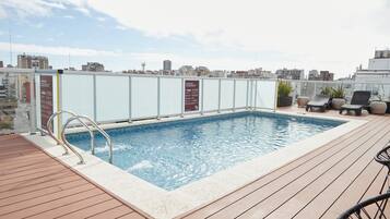 A rooftop pool with a wooden deck, lounge chairs, and a cityscape view.