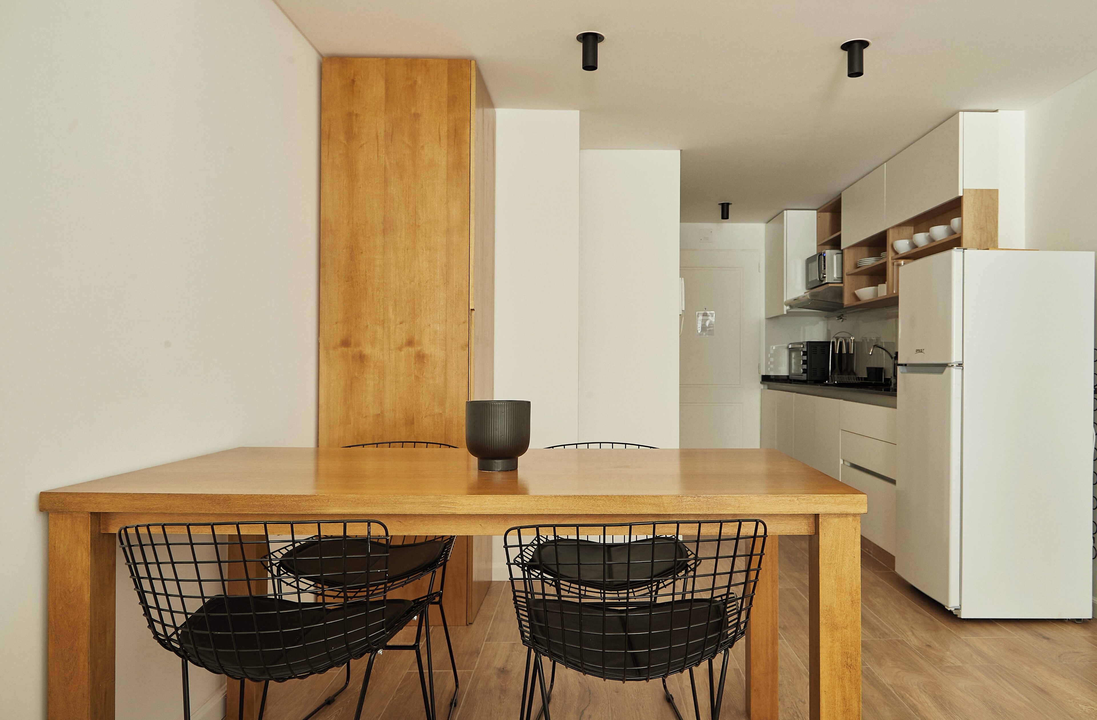 A modern kitchen with a wooden table and black wire chairs, white cabinets, and a white refrigerator.