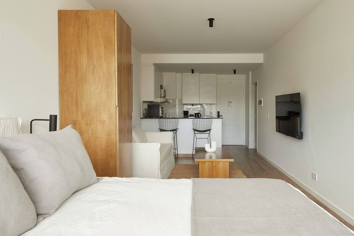 A modern hotel room with a large bed, a wooden wardrobe, a kitchenette, and a TV mounted on the wall.