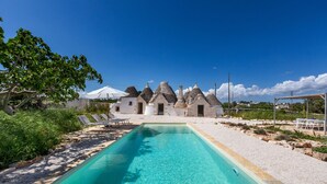 A swimming pool with a clear blue water, surrounded by white buildings with conical roofs, and a tree to the left. - 4003 Brunaldi Trulli by Perle di Puglia (Martina Franca)