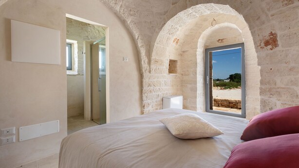 A bedroom with a stone wall, a bed with white and pink bedding, and a view of the beach through an arched window. - 4003 Brunaldi Trulli by Perle di Puglia (Martina Franca)