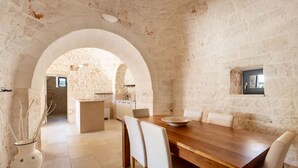 A dining area with a wooden table and white chairs, set against a stone wall with an arched doorway leading to a kitchen. - 4003 Brunaldi Trulli by Perle di Puglia (Martina Franca)