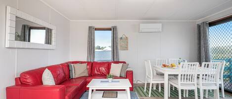A red leather sofa, white dining table, and chairs in a room with a large window, white walls, and a ceiling light.