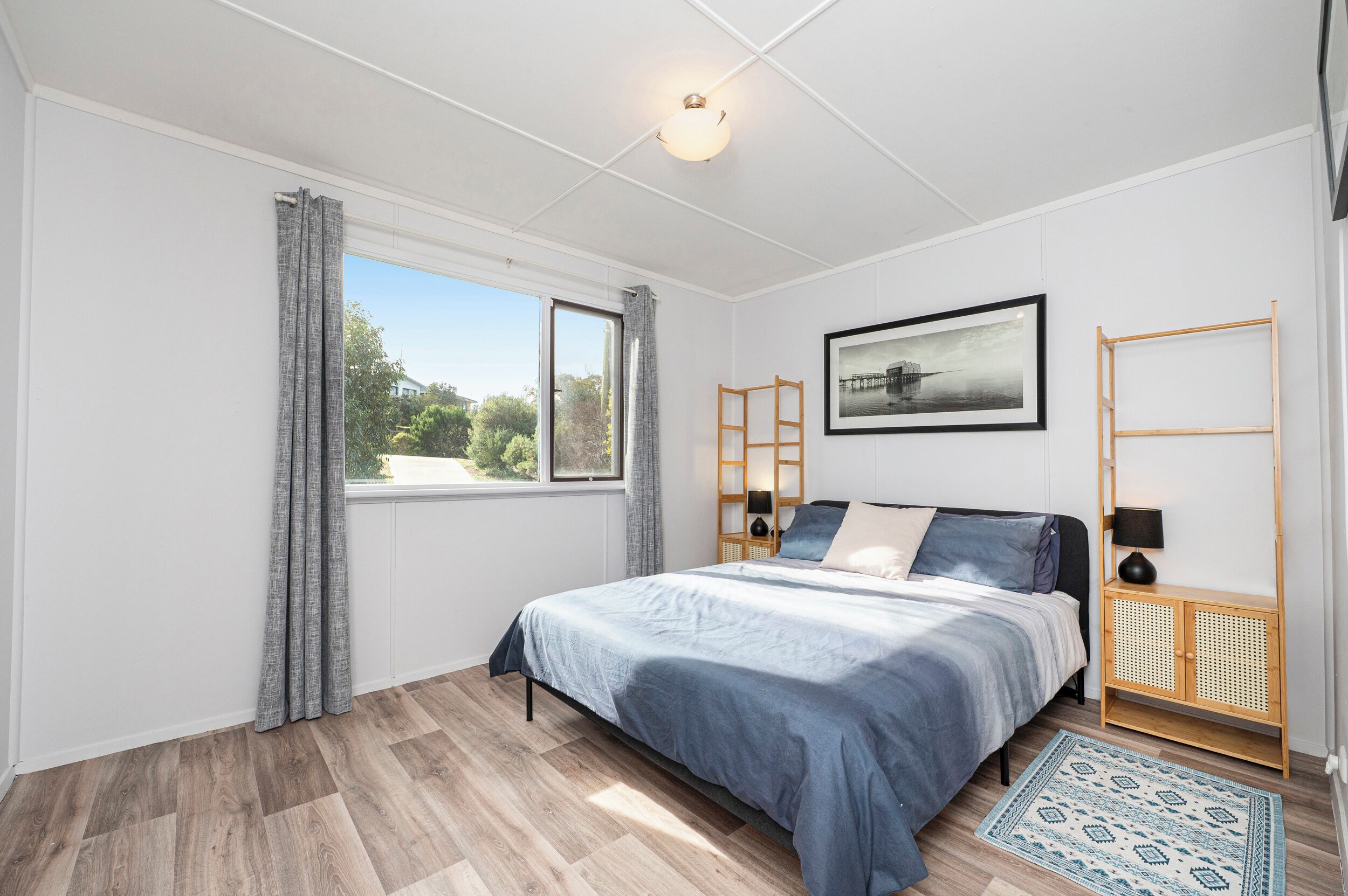 A bedroom with a bed, a window with curtains, a framed picture, a wooden ladder, a bedside table with a lamp, and a patterned rug.
