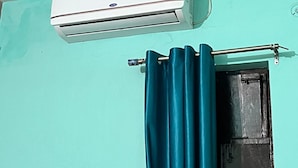 A room with a teal wall, a white air conditioning unit mounted above a door, and a teal curtain partially drawn. - Hotel Aryavart (Meerut)
