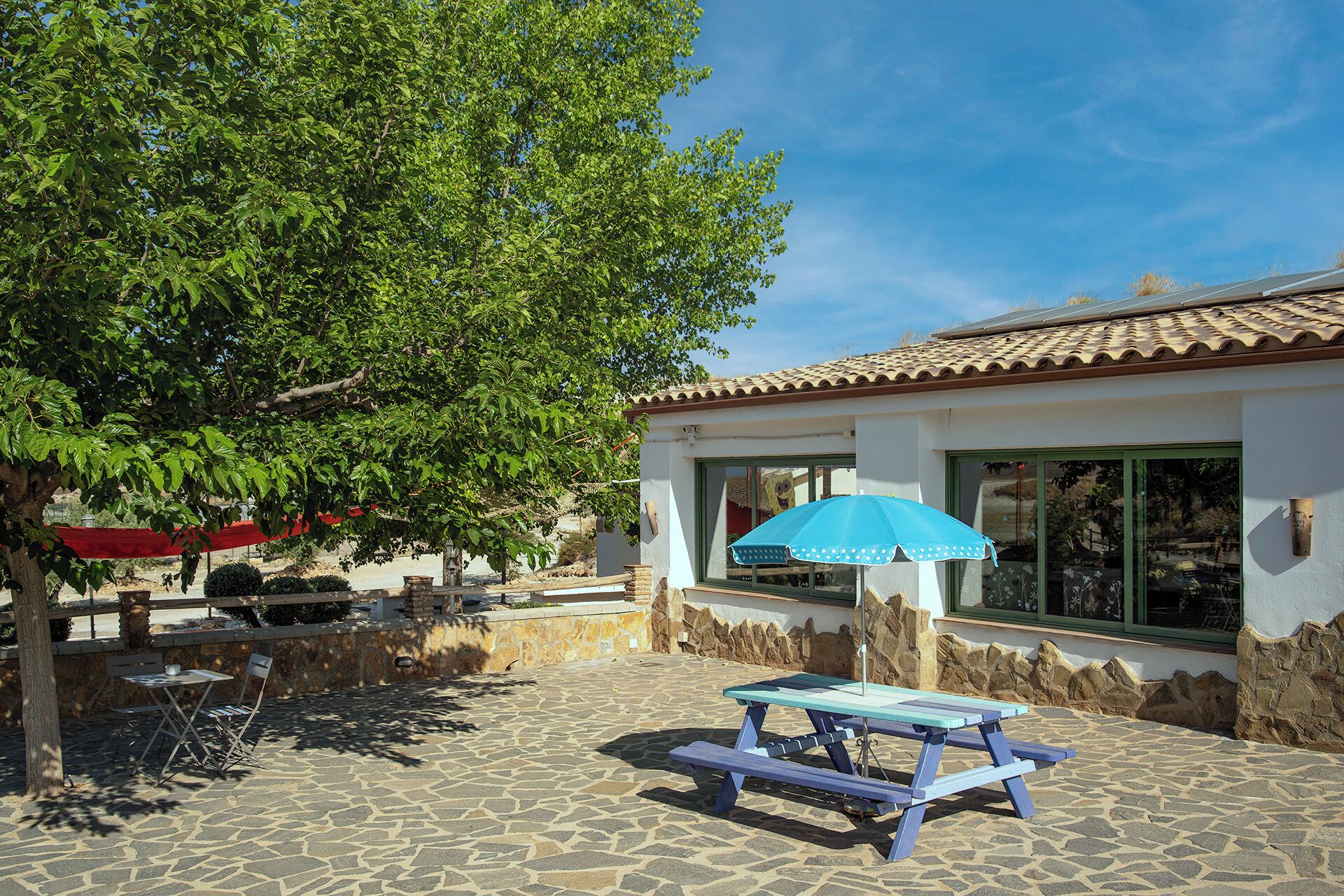 A patio with a picnic table and chairs, a stone floor, a building with a tiled roof, and a large tree.