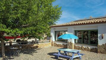 A patio with a picnic table and chairs, a stone floor, a building with a tiled roof, and a large tree.