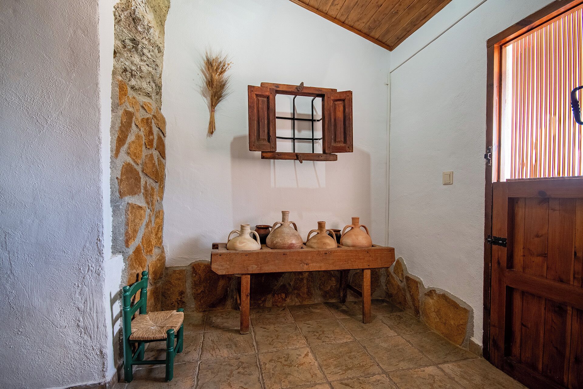 Una habitación rústica con pared de piedra, un banco de madera con cerámica, una silla verde y una puerta de madera.