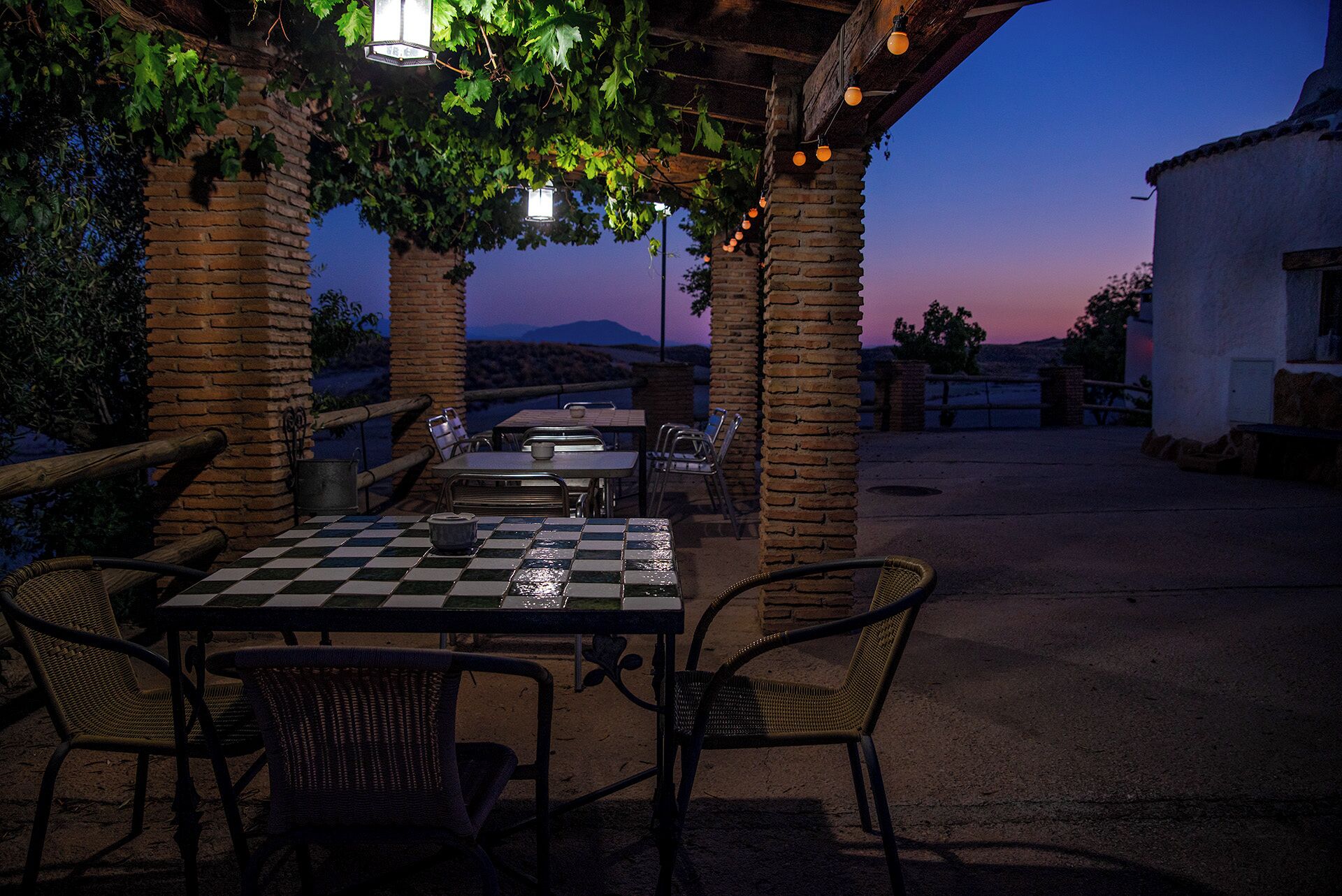 Un patio al aire libre con una mesa a cuadros y sillas de mimbre, rodeado de pilares de ladrillo y una pérgola con luces colgantes.