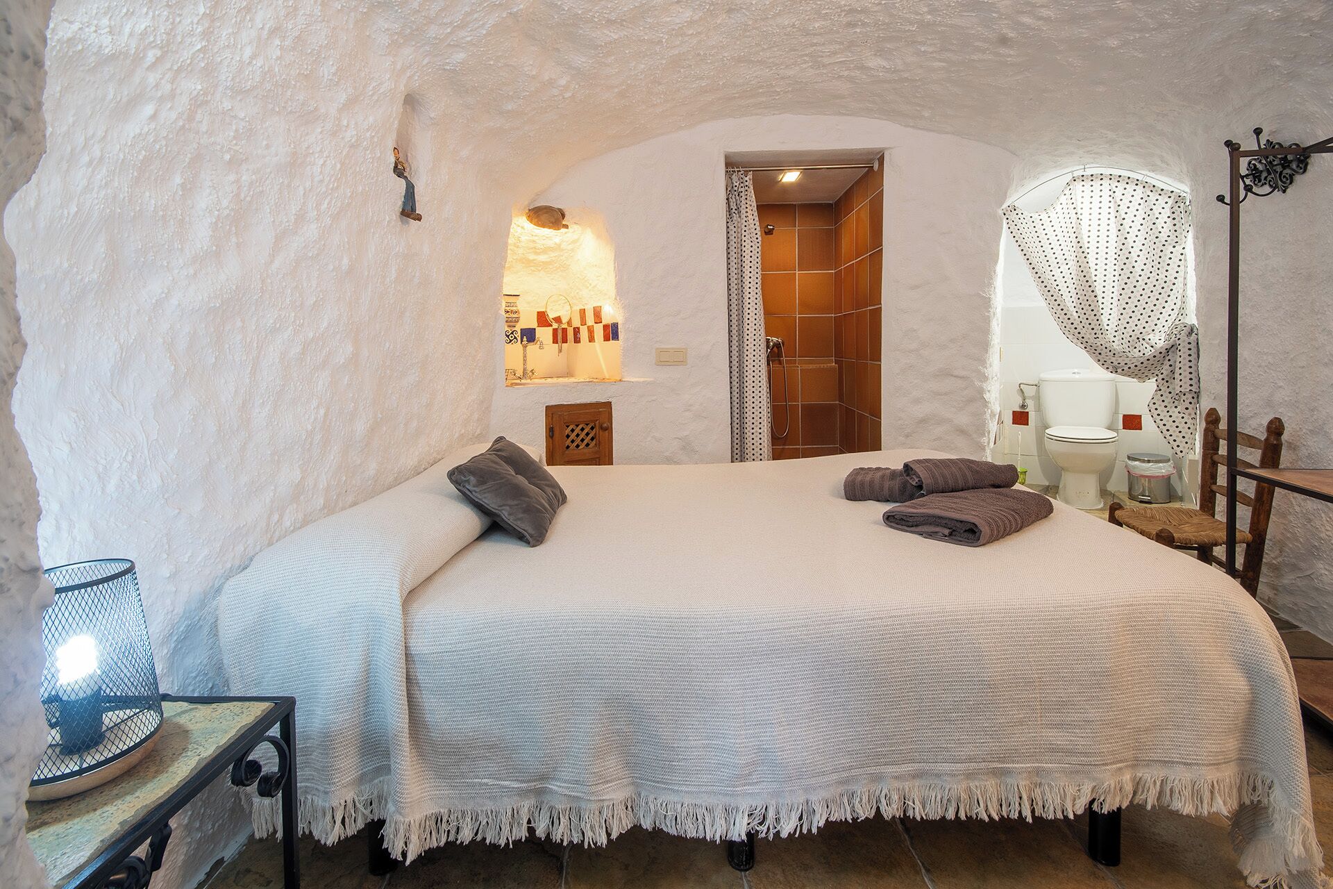 A cozy bedroom with a large bed, a wooden headboard, a small table with a lamp, and a bathroom visible through an archway.