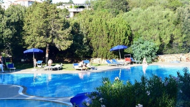 Una piscina con sillones de descanso y sombrillas, rodeada de árboles y un muro de piedra.