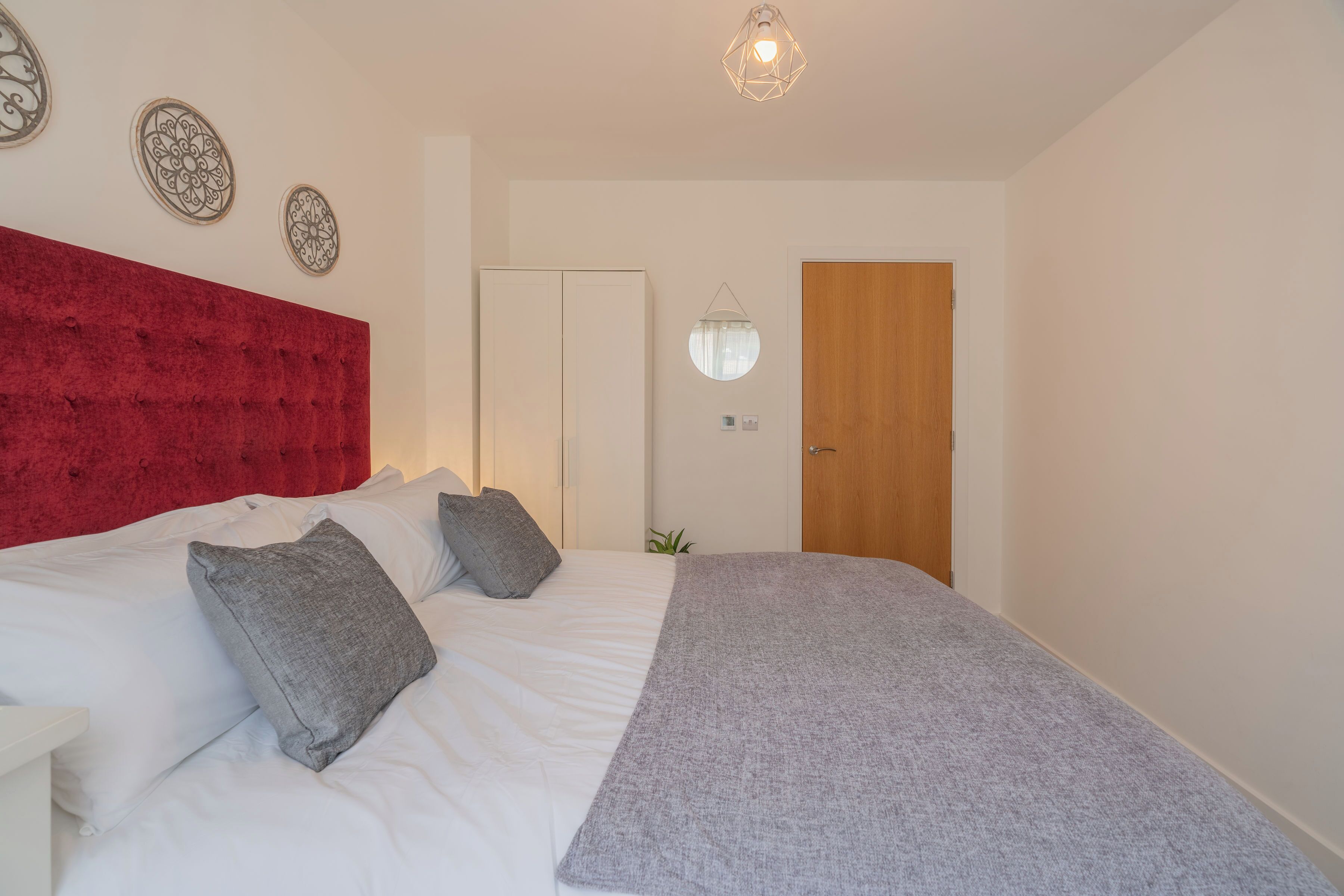A bedroom with a red headboard, a bed with white bedding, a grey throw blanket, and a wooden door.
