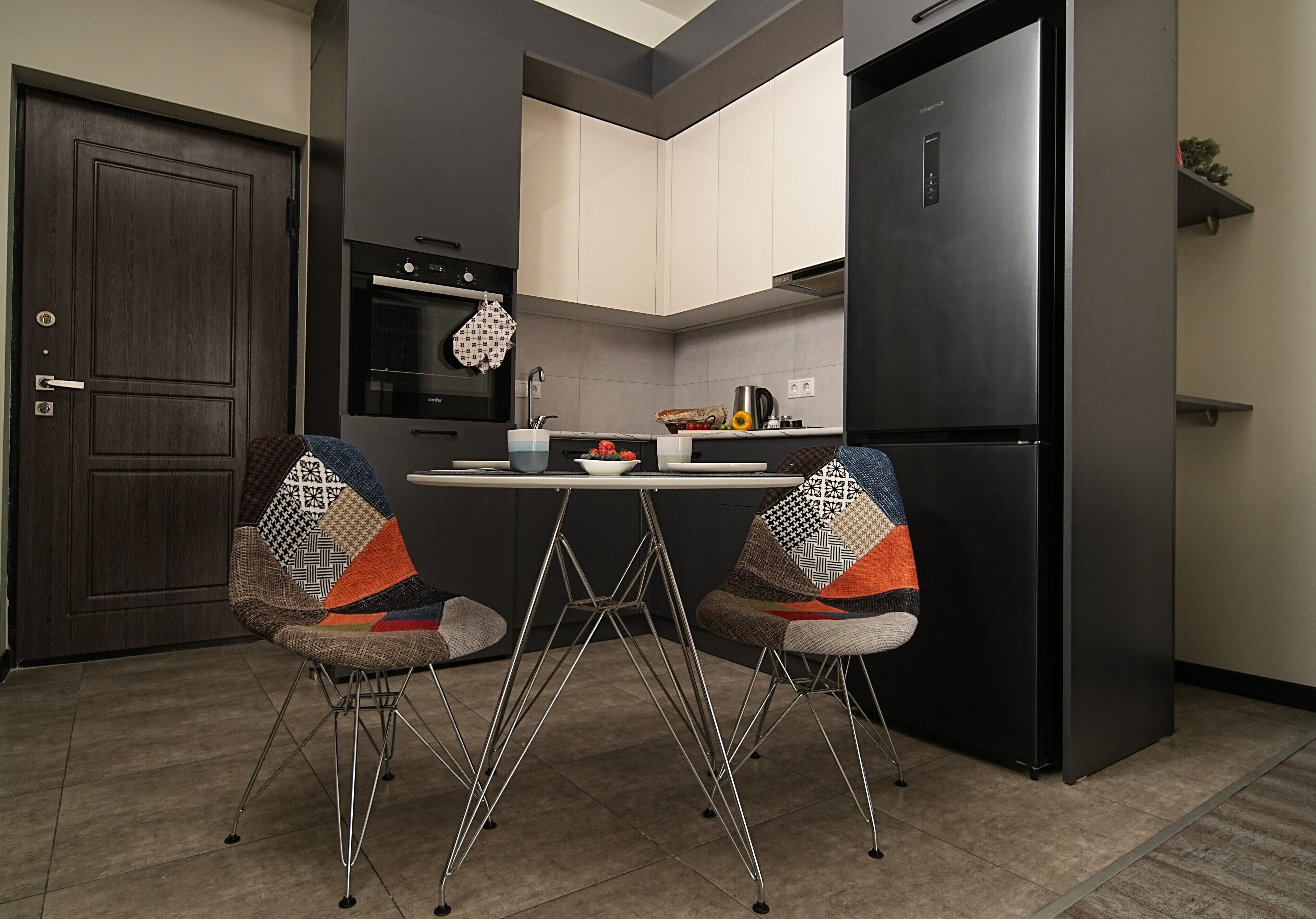 Una cocina moderna con un refrigerador negro, dos sillas estampadas, una mesa de comedor con un cuenco de fruta y un guante de cocina estampado.
