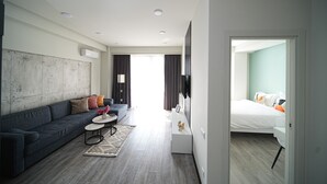 A modern hotel room with a grey sofa, a small round table, a flat-screen TV mounted on the wall, and a bed with white bedding. - Modern 1-bedroom apartment by Sweet Home (Yerevan)