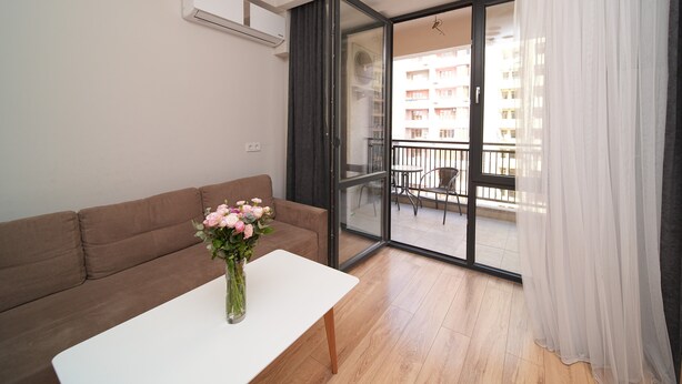 A modern living room with a brown sofa, a white coffee table, and a vase of flowers. There is a sliding glass door leading to a balcony with a view of a building. - Modern 1-bedroom apartment by Sweet Home (Yerevan)