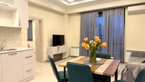 A modern kitchen with white cabinets, a wooden countertop, and a sink. There is a dining area with a wooden table, green upholstered chairs, and a vase with flowers. A television is on a white cabinet, and there is a window with curtains. - Modern 1-bedroom apartment by Sweet Home (Yerevan)