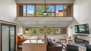 A spacious living area with a large TV, a fireplace, and multiple windows offering views of the outdoors. - Cliffside Suite | AC, Pool, Hot Tub, Golf & Ski (Durango)
