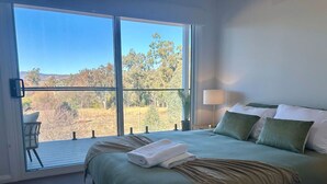 A bedroom with a large window, a bed with green bedding, a bedside table with a lamp, and a view of trees and hills. - House on the hill | Kangaroo lookout (Wodonga)