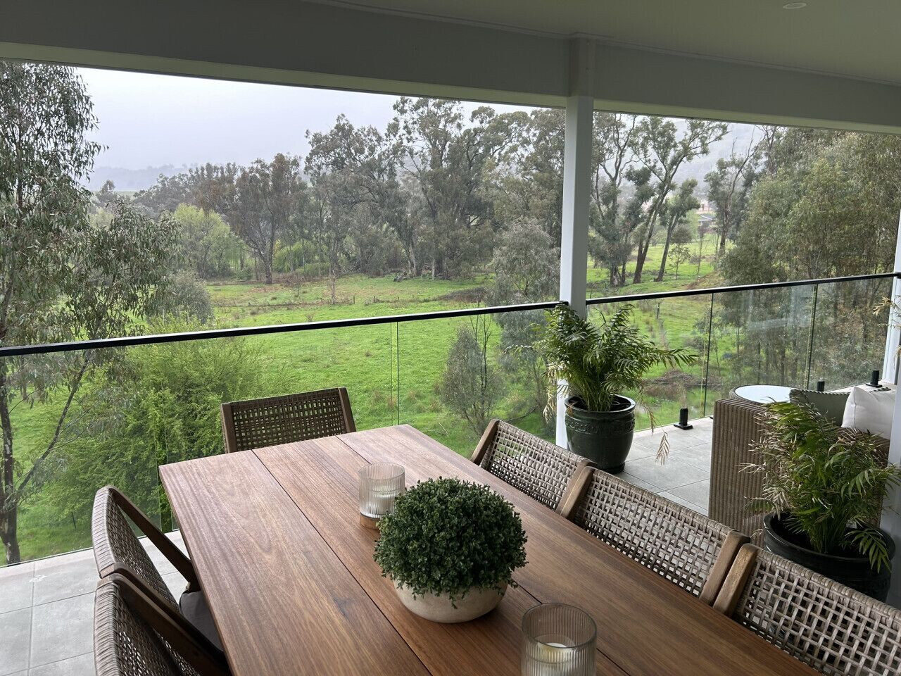 Un coin repas avec une table et des chaises en bois, des plantes en pot et une vue sur un paysage verdoyant grâce à de grandes fenêtres.