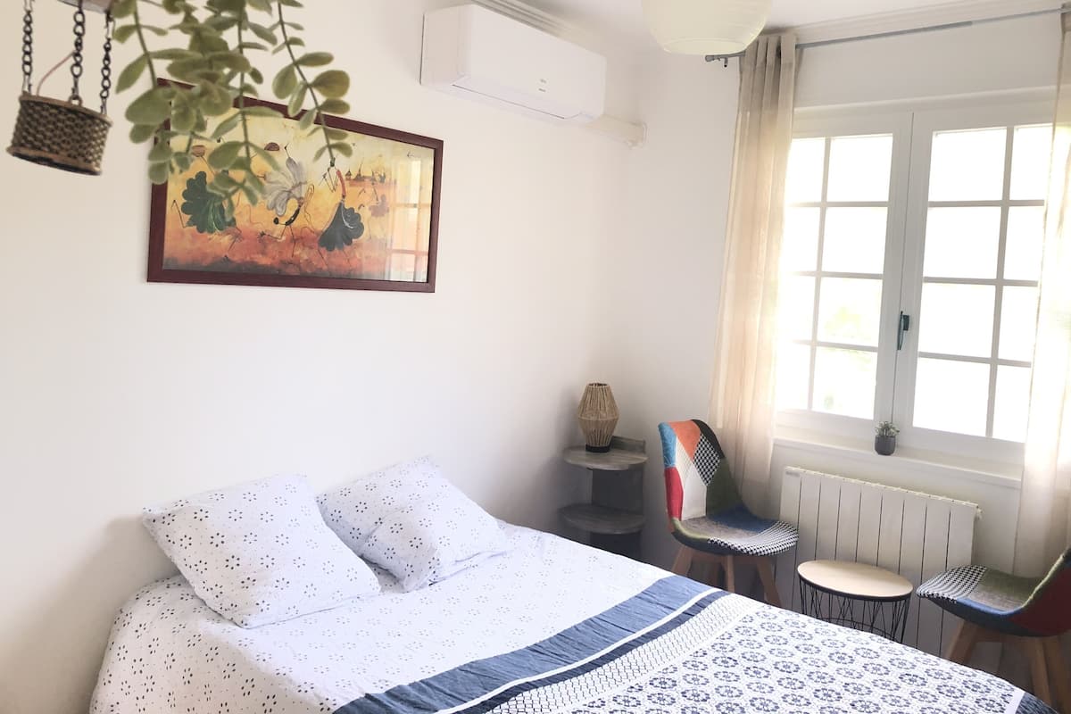 A bedroom with a bed, a chair, a small table, a window with curtains, and a hanging plant.
