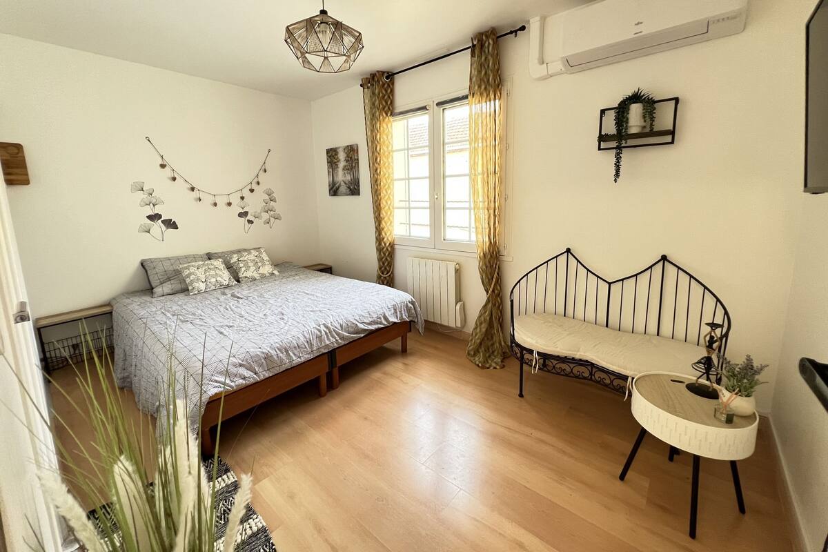 A bedroom with a bed, a small bedside table, a chair, a window with curtains, a radiator, and a wall-mounted shelf.