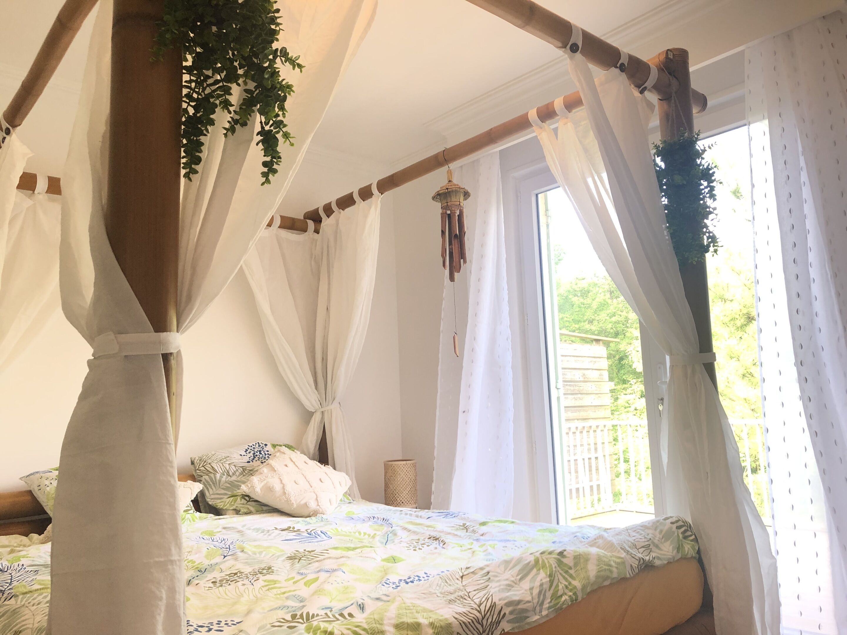A bedroom with a canopy bed, white curtains, and a view of greenery outside.