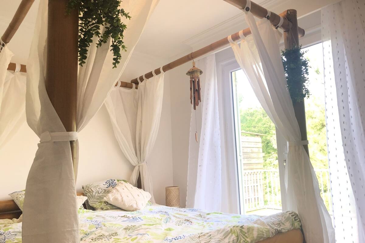 A bedroom with a canopy bed, white curtains, and a view of greenery outside.