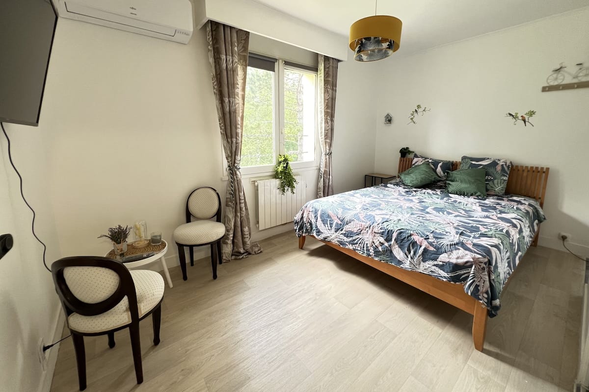 A bedroom with a bed, a chair, a small table, a window with curtains, a ceiling fan, and wall decorations.
