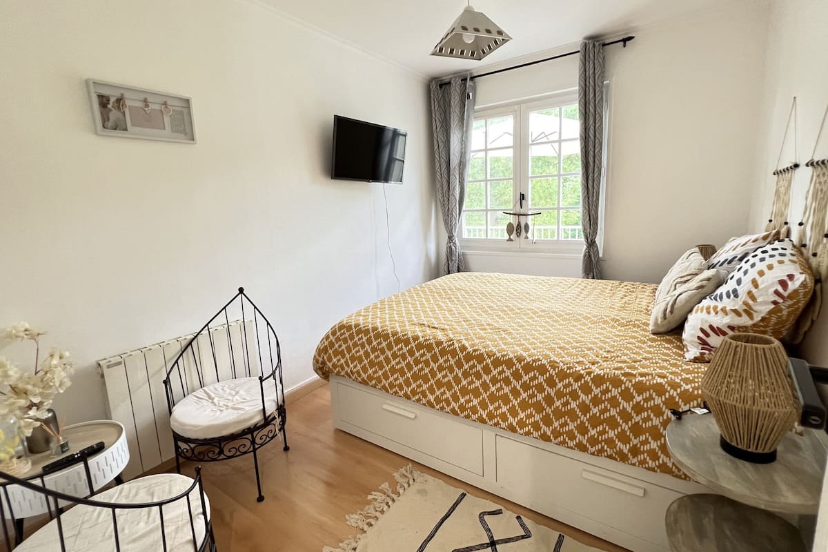 A bedroom with a bed, a TV, a window with curtains, a chair, and a bedside table with a lamp.