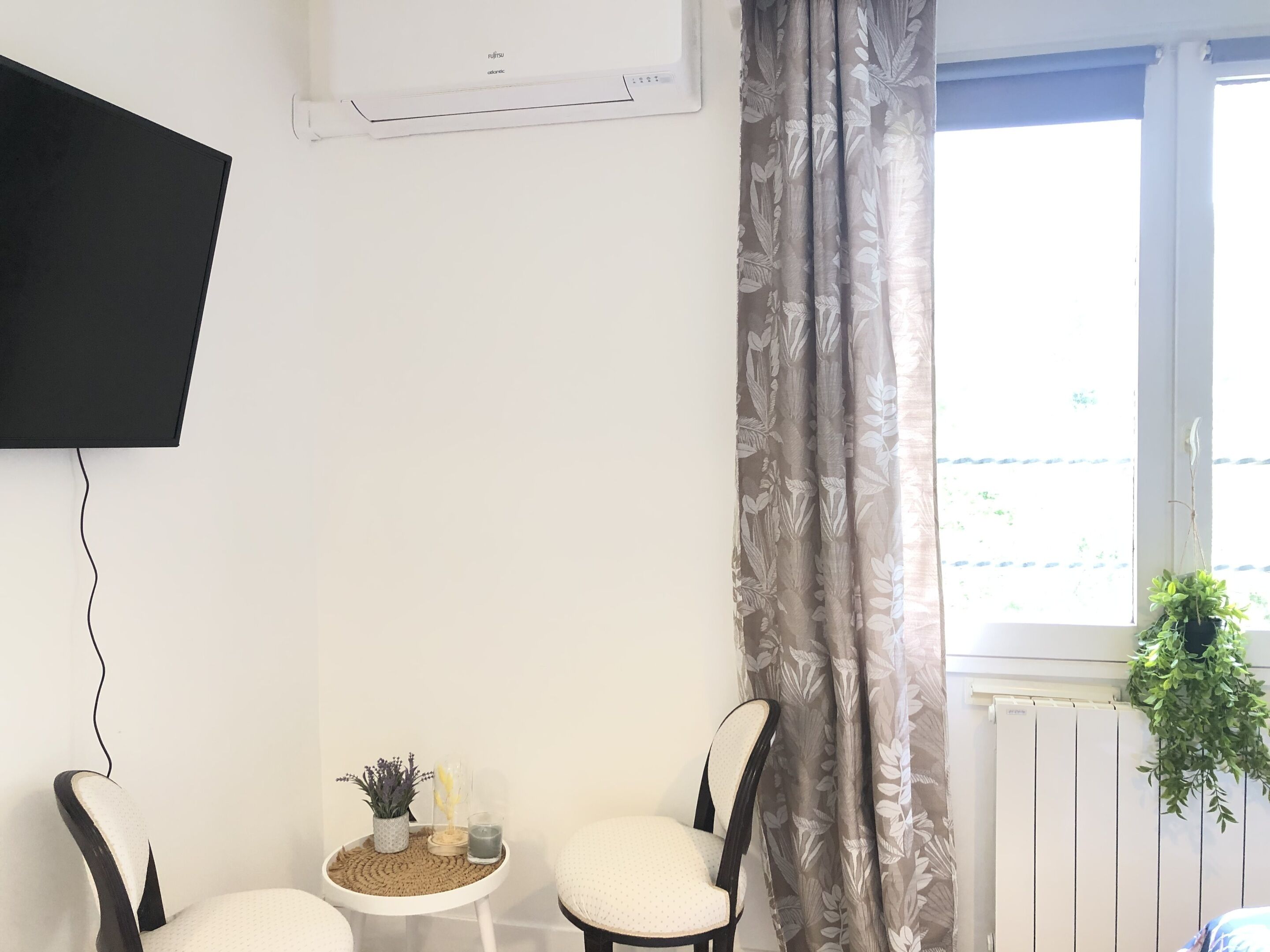 Una habitación con una televisión de pantalla plana en la pared, dos sillas, una mesita con objetos decorativos y una ventana con cortinas.