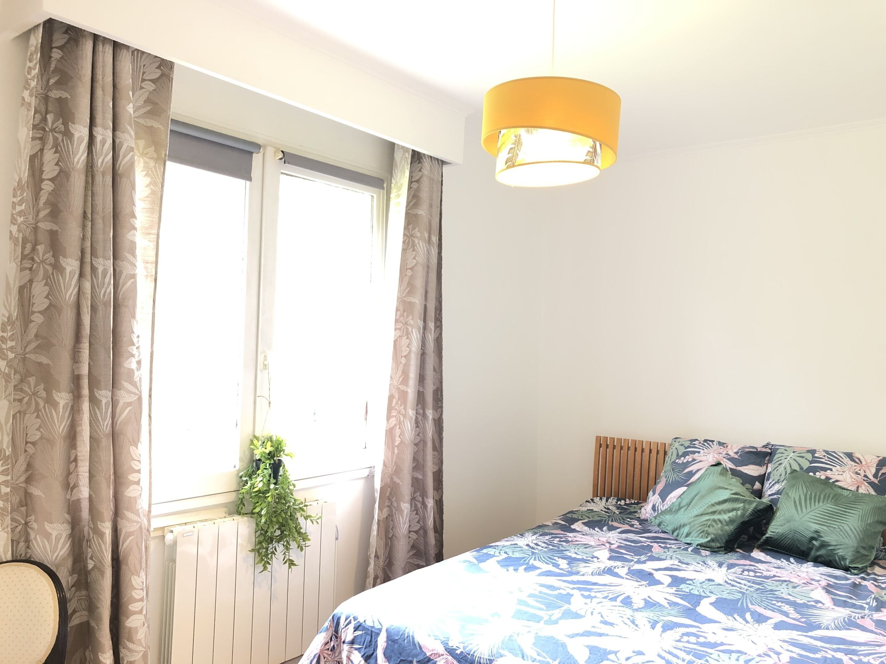 Een slaapkamer met een bed, gordijnen, een hangende plant en een plafondlamp.
