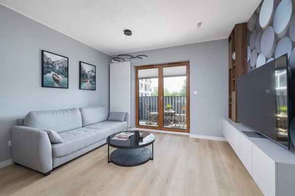 Una sala de estar moderna con un sofá gris, una mesa redonda de centro y un televisor de pantalla plana sobre un mueble blanco. Hay dos cuadros en la pared y un ventanal corredero que da a un espacio exterior.