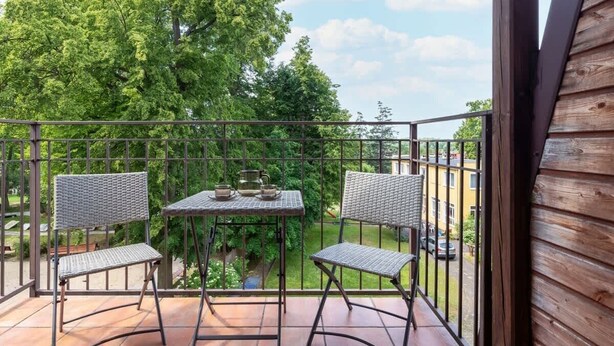 A balcony with two wicker chairs and a small table, overlooking a green garden and a building.