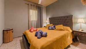 Un dormitori amb llit, ventilador de sostre, tauleta de nit amb làmpada i finestra amb cortines.