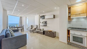 A modern living room with a grey sofa, a dining area with chairs, a kitchen with wooden cabinets and a stove, and a view of the sea through large windows. - Las Americas Ocean view Sunsets (Arona)