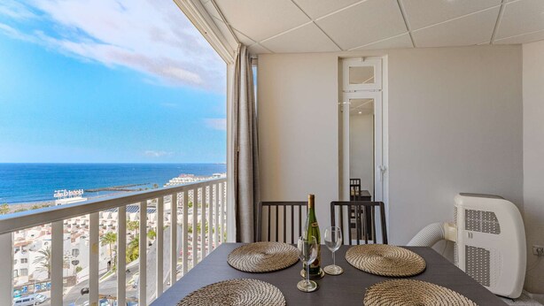 A balcony with a view of the sea, a table with woven placemats, a bottle of wine, and a fan. - Las Americas Ocean view Sunsets (Arona)