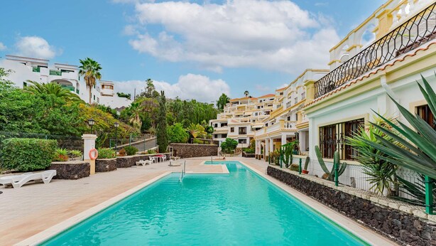 A swimming pool with a clear blue water, surrounded by a white building with balconies, and lush greenery in the background.