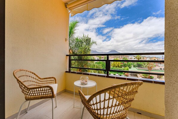 A balcony with two wicker chairs and a small table, overlooking a cityscape with greenery and buildings.