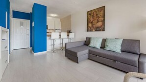 En moderne stue med en grå hjørnesofa, en hvid dør og et køkken med hvide skabe samt en blå accentvæg.