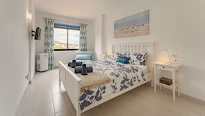 Et soveværelse med en seng, et natbord, en lampe, et vindue med gardiner og et billede på væggen.